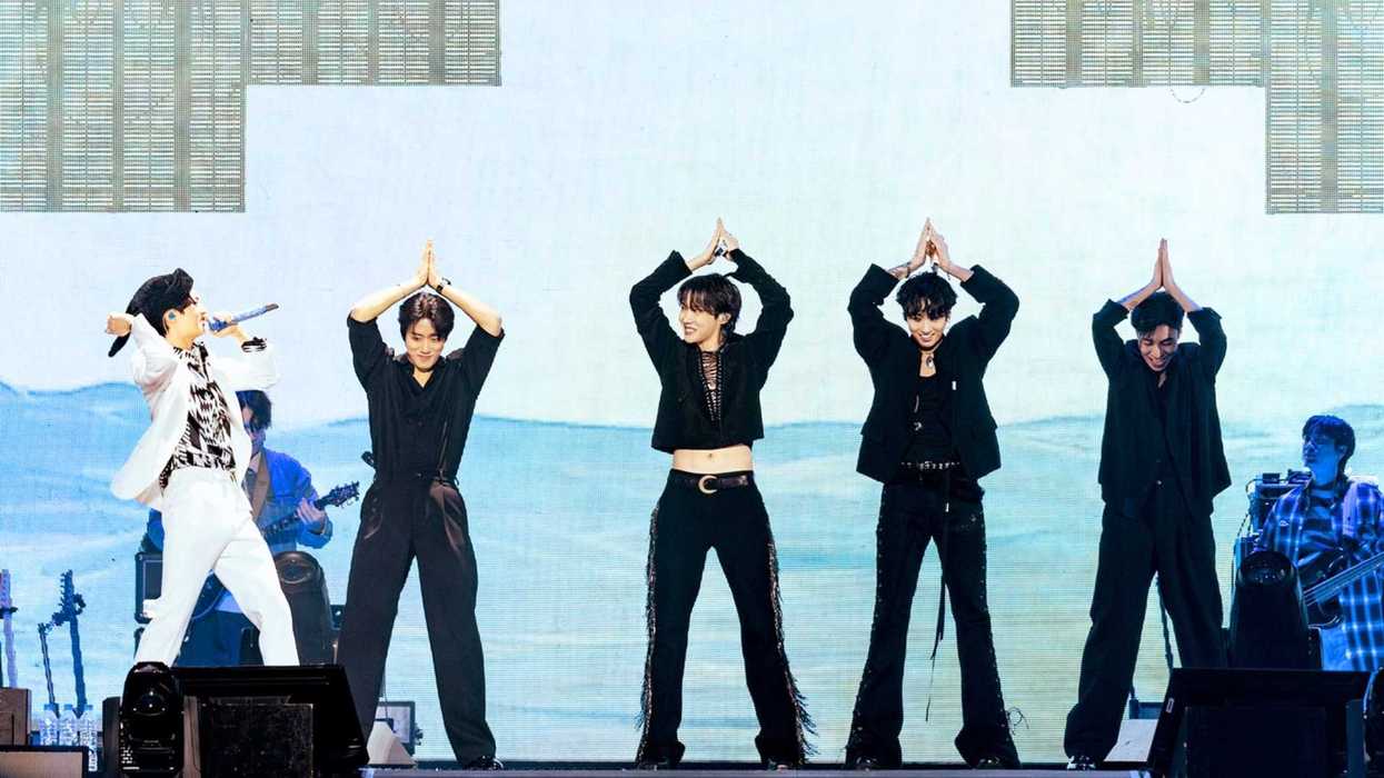 Five performers dance onstage, one singing with a microphone, colorful backdrop.