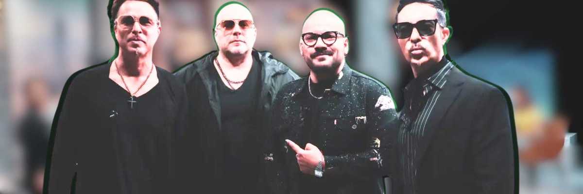 Four men in black outfits and sunglasses pose together indoors.