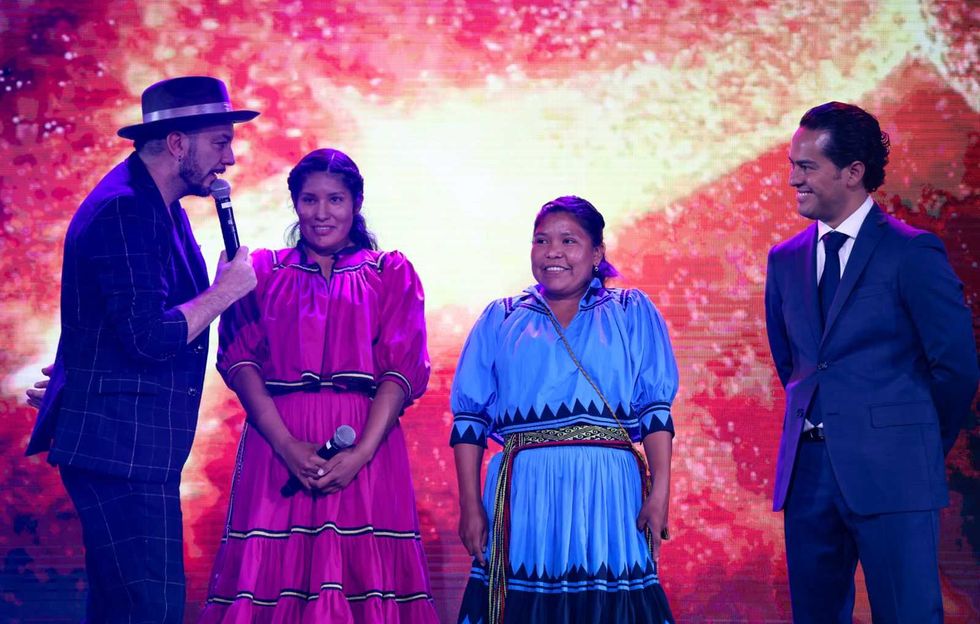 Four people on stage, two women in colorful dresses, two men in suits, under bright lights.