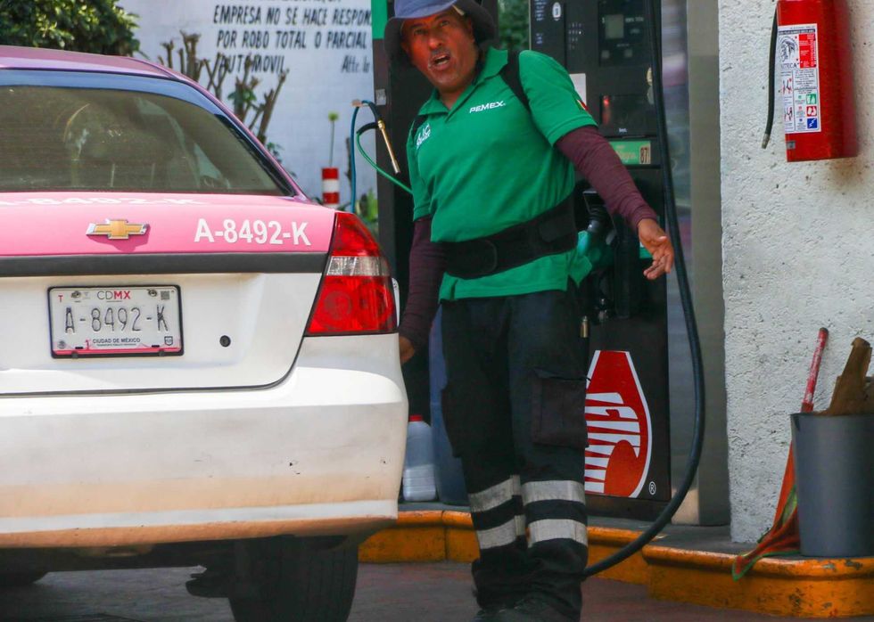 Gas station attendant fueling a taxi, wearing a green shirt and hat, next to a dispenser.