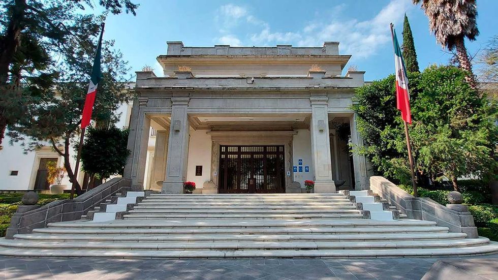 Grand entrance with steps, columns, and two Mexican flags, flanked by lush greenery.