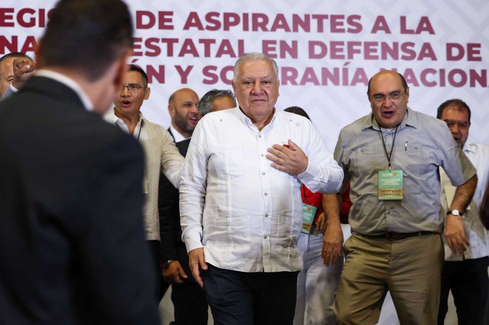 Group of men at a formal event, one holding his chest, with a banner in the background.