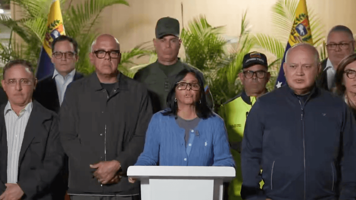 Group of officials standing, one speaking at a podium with flags in the background.