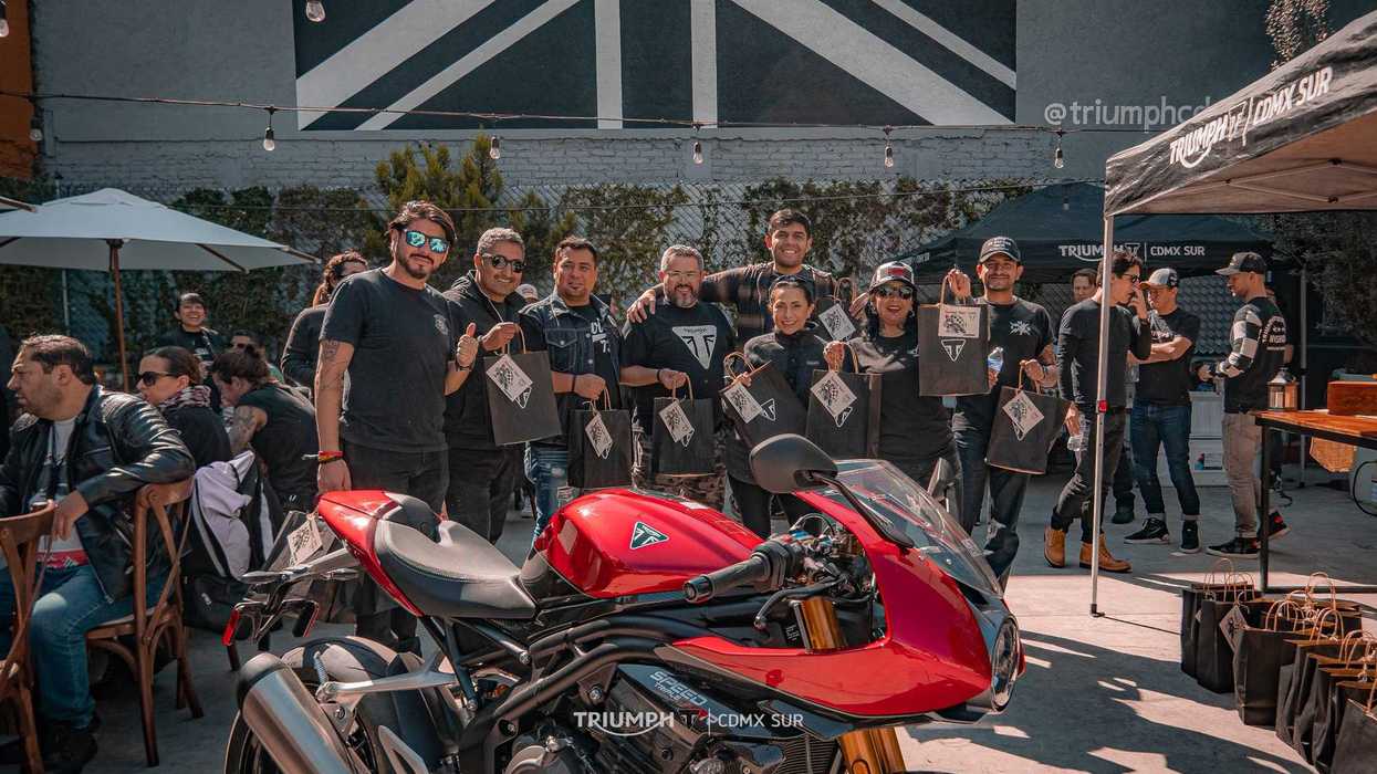 Group posing behind a red motorcycle at a Triumph event, with branded bags in hand.