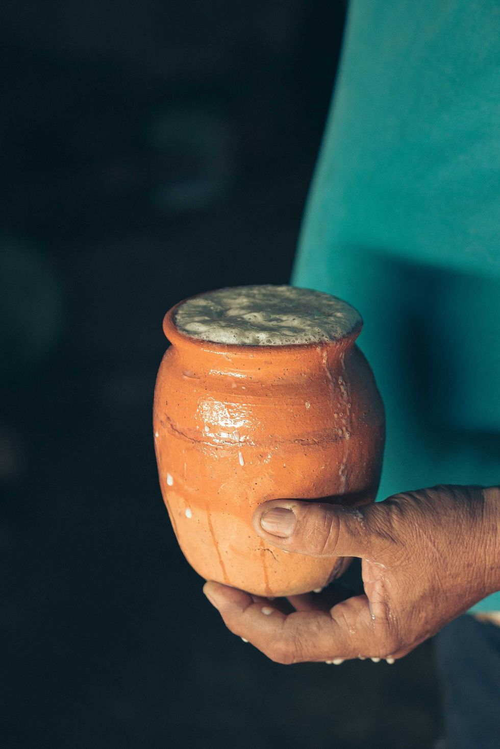 Hand holding a frothy drink in a clay pot against a dark background.