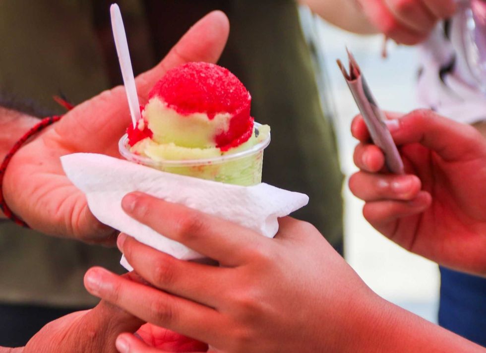 Hands exchanging a cup of colorful shaved ice with a spoon.