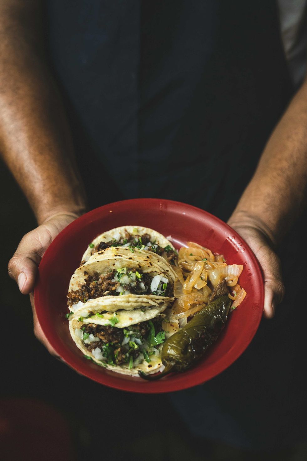 Hands holding a red plate with tacos, onions, and a jalape\u00f1o.