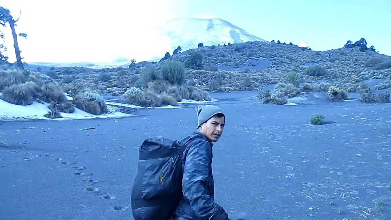 Hiker in gray beanie trekking across a volcanic landscape towards a snowy mountain.
