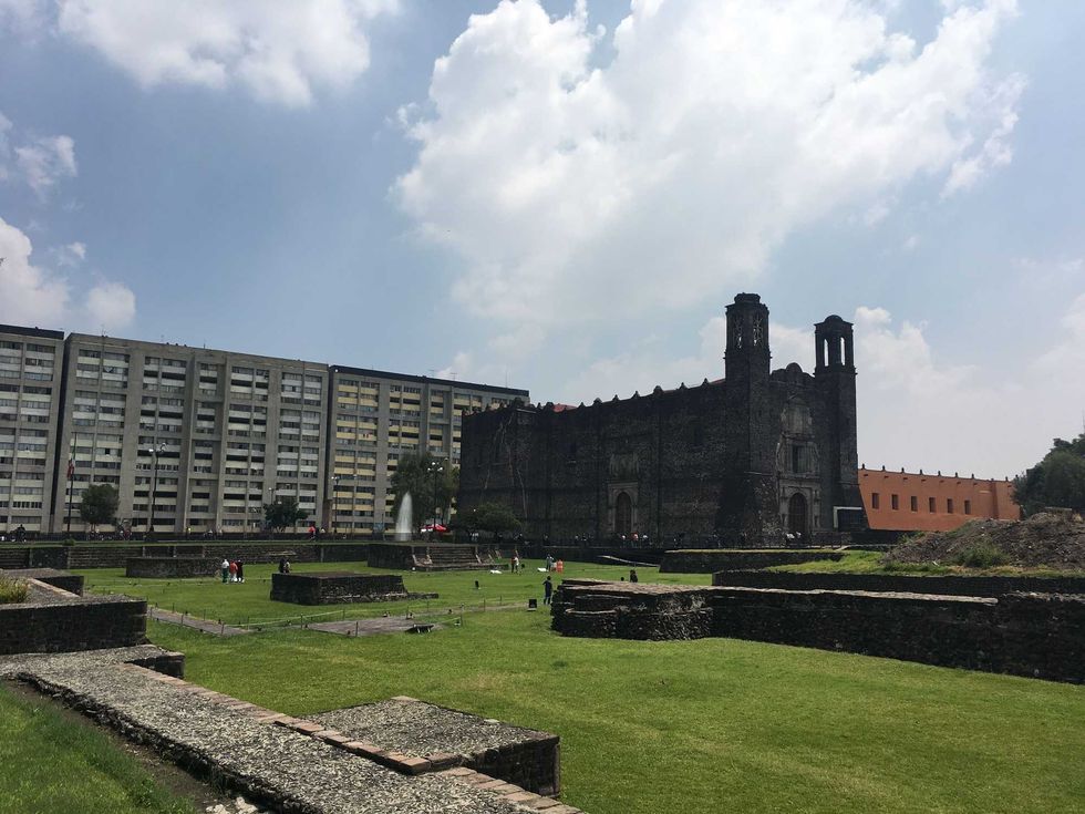Historic stone church next to modern buildings under a cloudy sky.