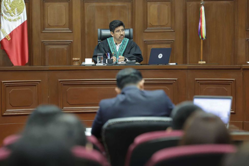 Judge presides over court session, wearing robe, with flags behind.