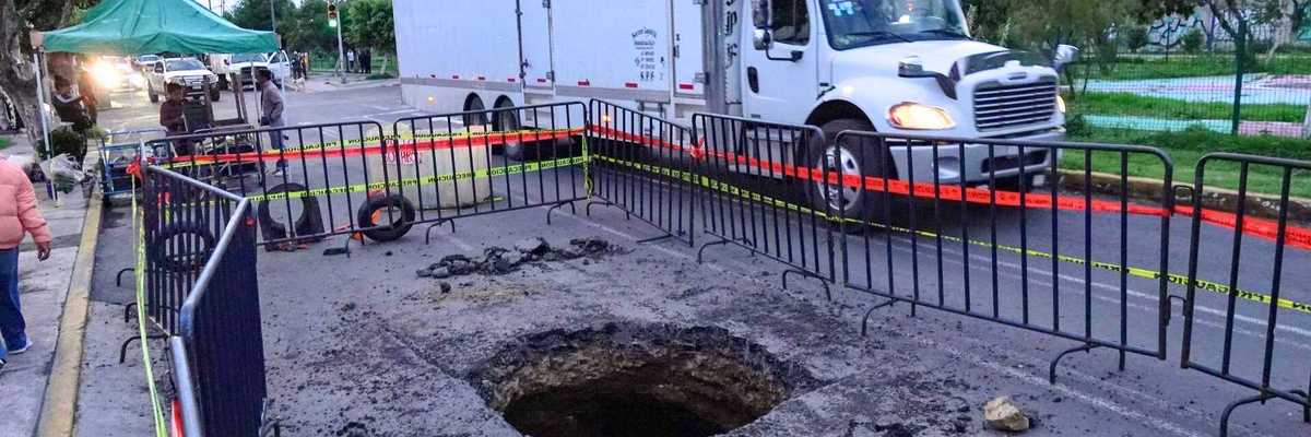 Large sinkhole on road, fenced for safety; truck passing by, traffic light visible.