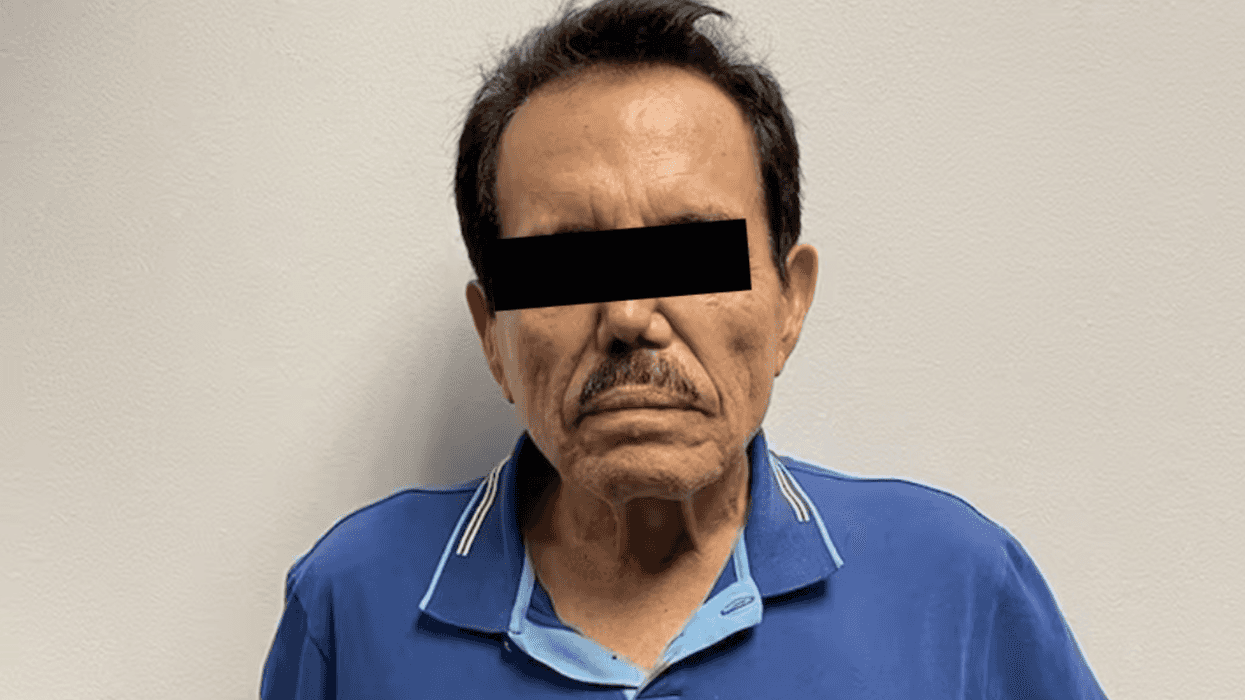 Man in a blue shirt with black bar obscuring eyes, standing against a plain wall.