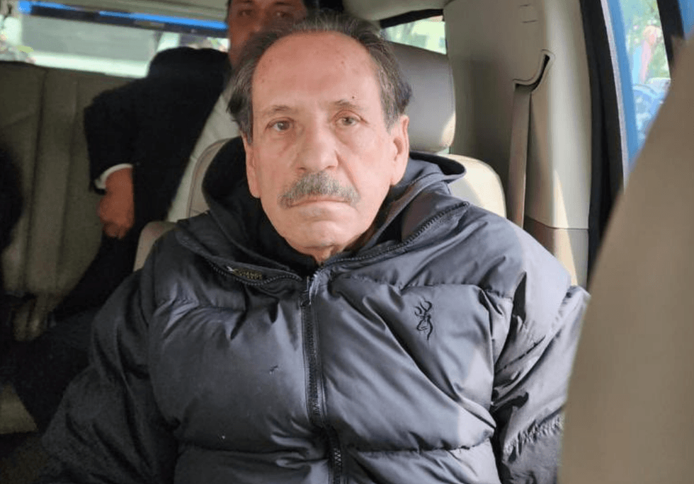 Man in a car wearing a black jacket, looking directly at the camera.