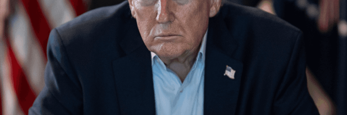 Man in a suit sitting with clasped hands, U.S. flags in the background.