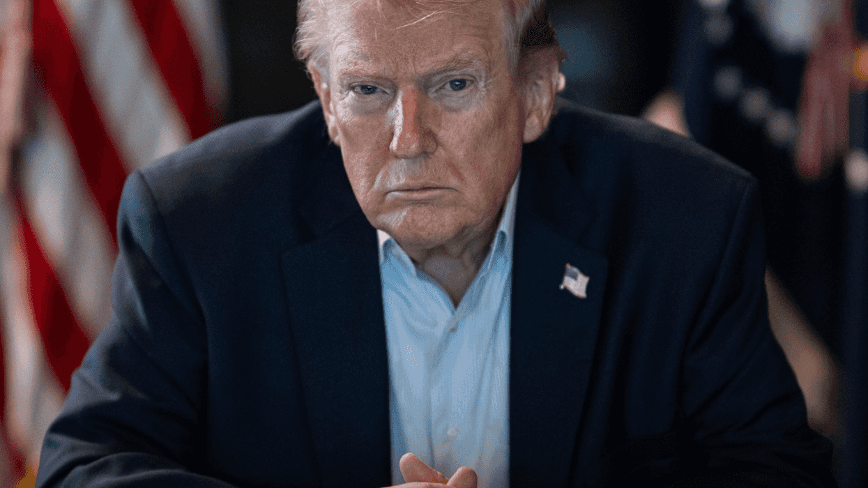 Man in a suit sitting with clasped hands, U.S. flags in the background.
