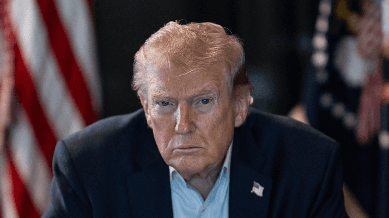 Man in a suit sitting with clasped hands, U.S. flags in the background.