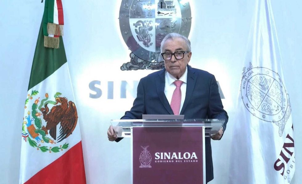 Man in a suit speaking at a lectern with Sinaloa flags and seal in the background.