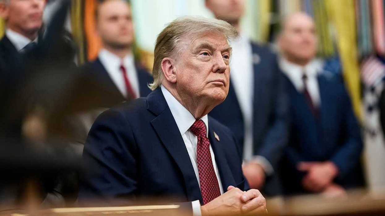 Man in a suit with hands clasped, surrounded by blurred figures in a formal setting.