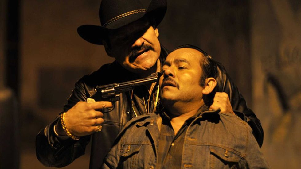 Man in black hat holds gun to another man's head in a dimly lit scene.