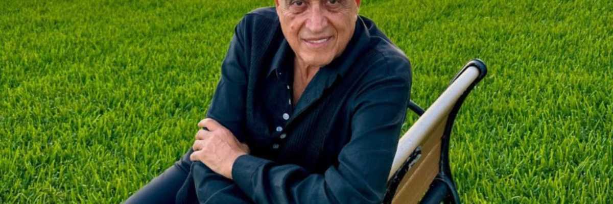 Man in black shirt sitting on a bench in a grassy area by the water.