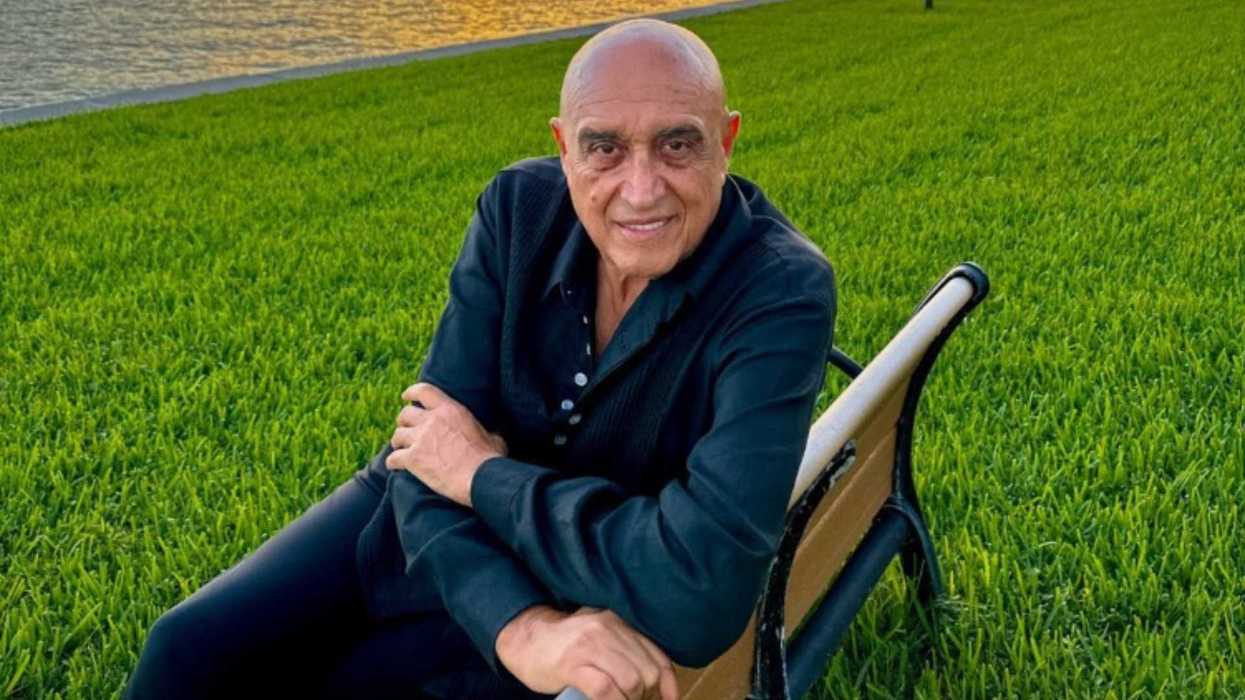 Man in black shirt sitting on a bench in a grassy area by the water.