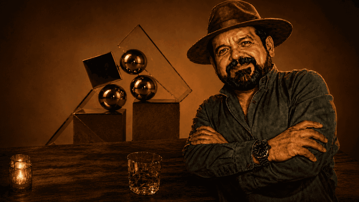 Man in hat with folded arms beside geometric sculptures and drink on a wooden table.
