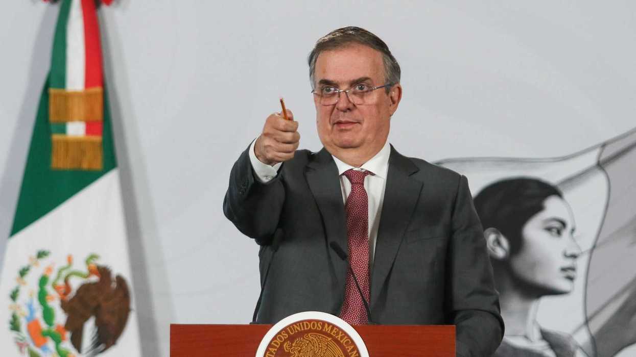 Man in suit gestures at podium with Mexican flag and mural in background.