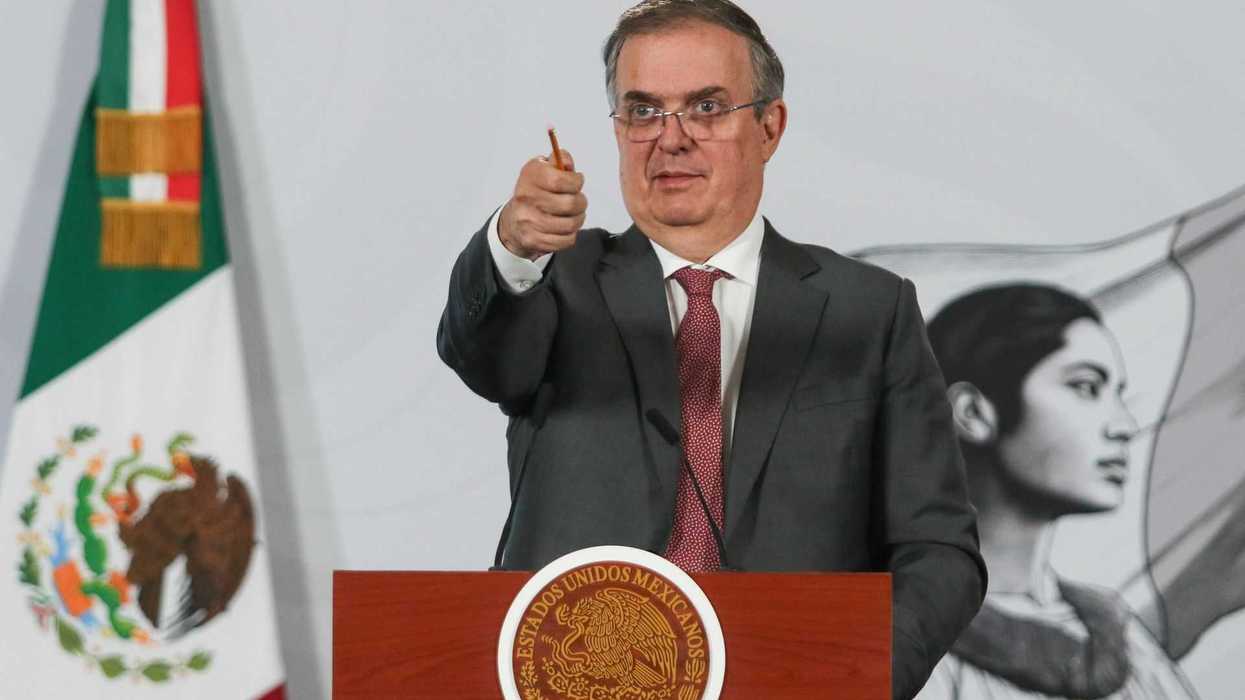 Man in suit gestures at podium with Mexican flag and mural in background.