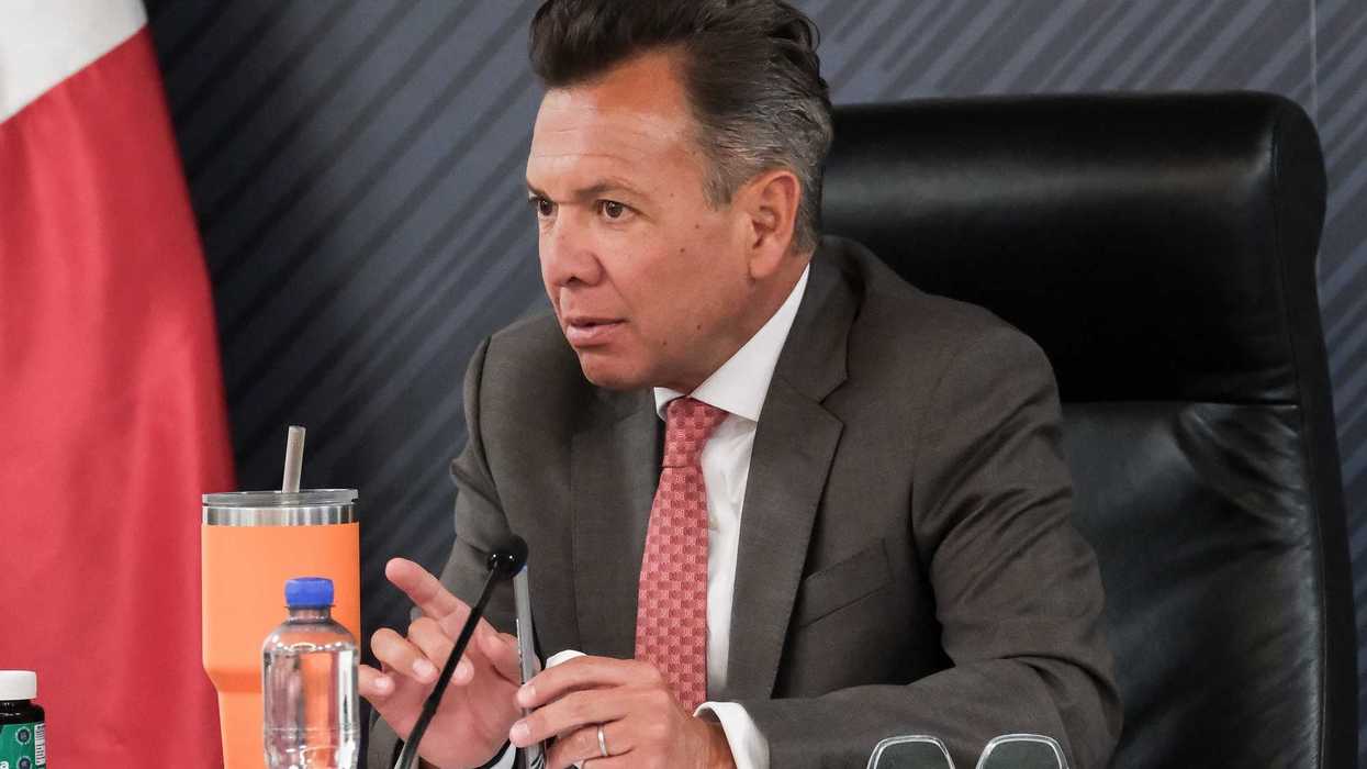 Man in suit speaking at a desk with a microphone and papers.