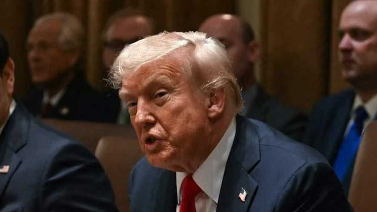 Man in suit speaking at a meeting, surrounded by other seated individuals.