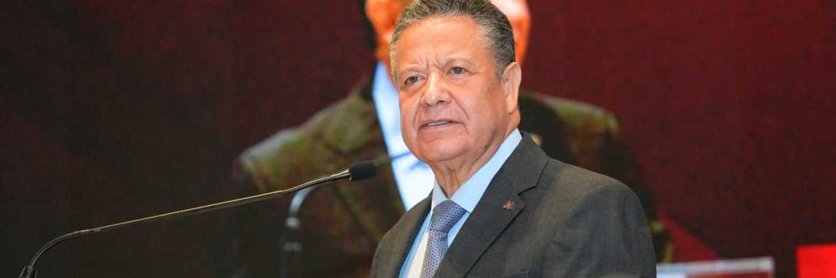Man in suit speaking at a podium with a large screen behind him.