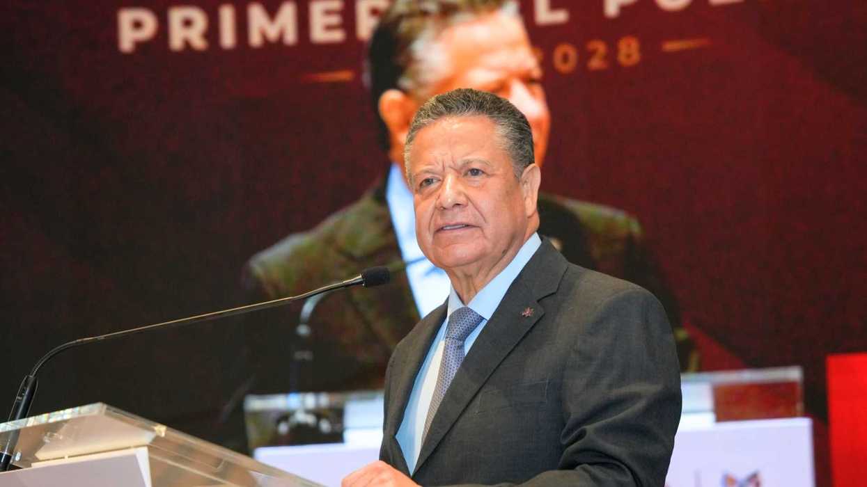 Man in suit speaking at a podium with a large screen behind him.