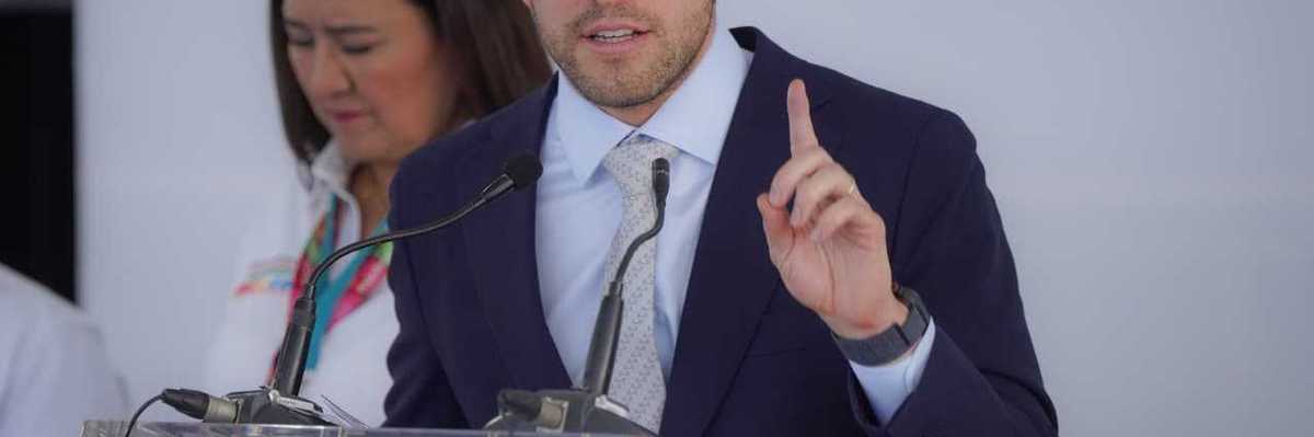 Man in suit speaking at a podium with a woman in background.
