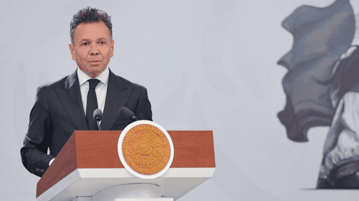 Man in suit speaks at podium with Mexican seal, white background.
