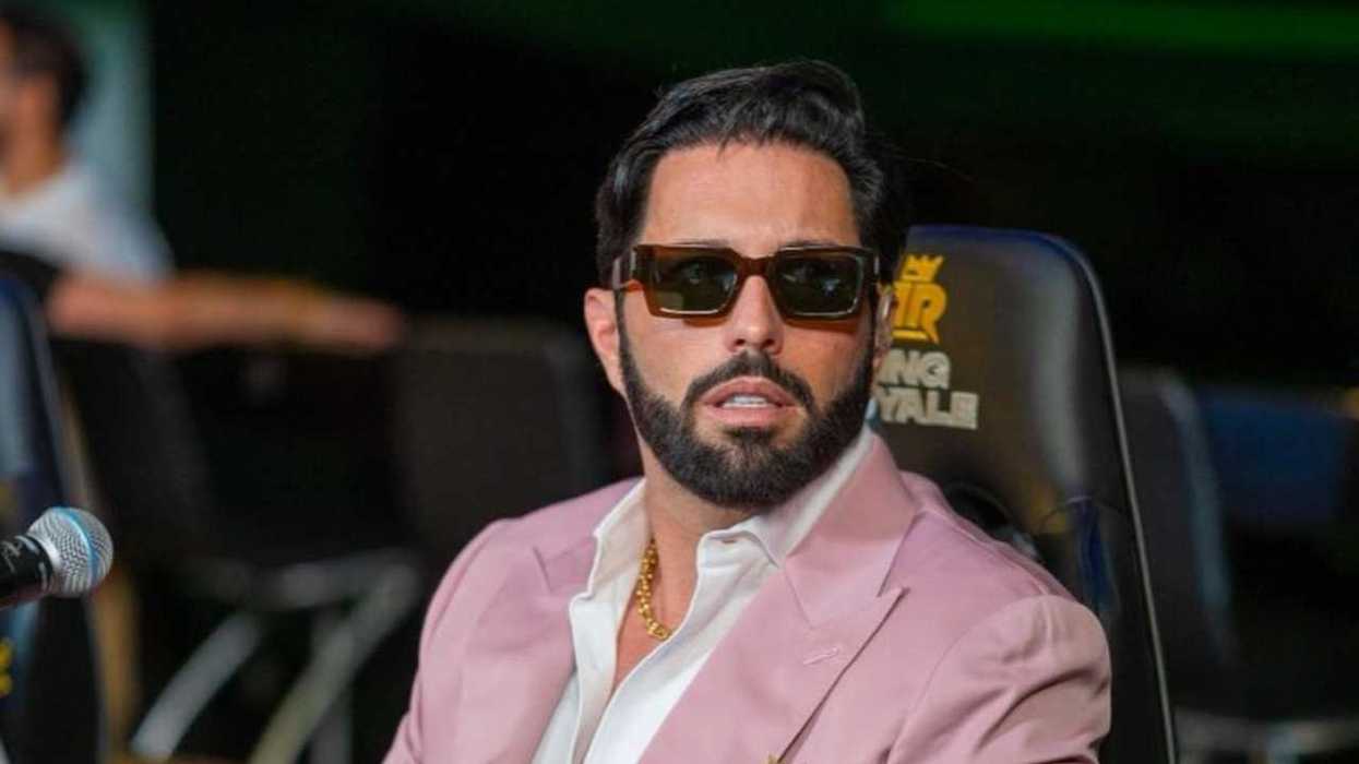 Man in sunglasses and pink blazer seated, microphone in view.