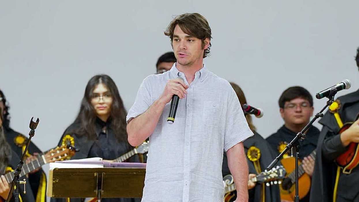 Man in white shirt speaking into a microphone on stage with musicians in the background.