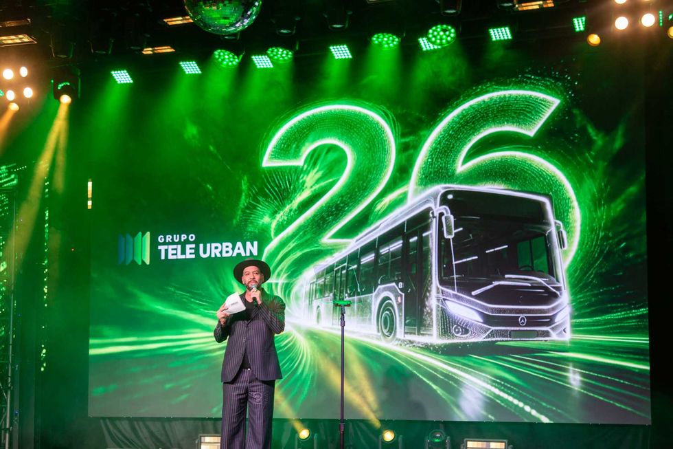 Man on stage with microphone, green bus backdrop, reads "26" and "Grupo Tele Urban."