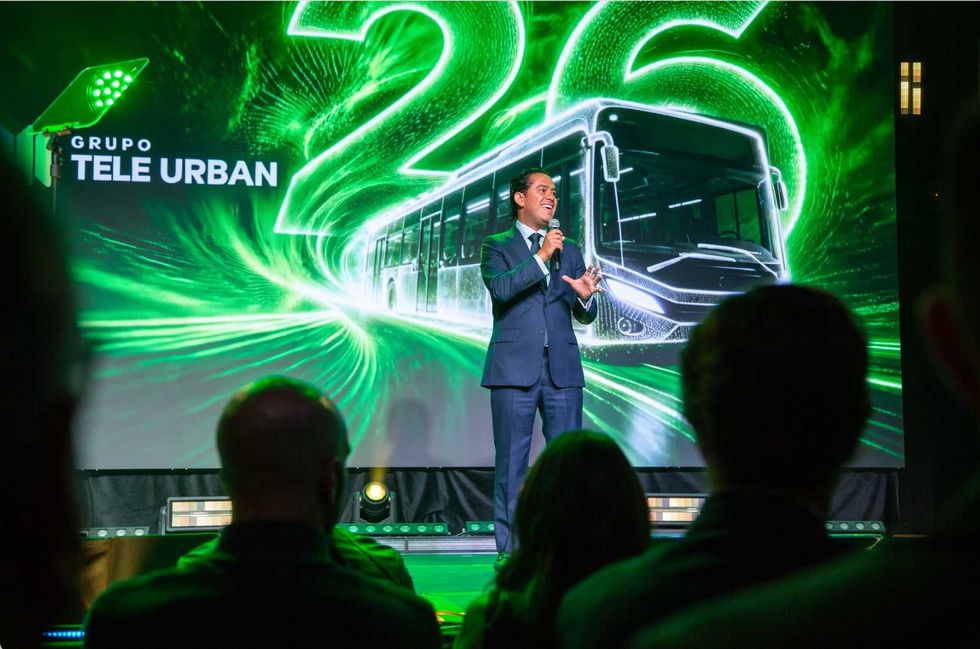 Man presenting on stage with "Grupo Tele Urban" backdrop featuring a green bus graphic.