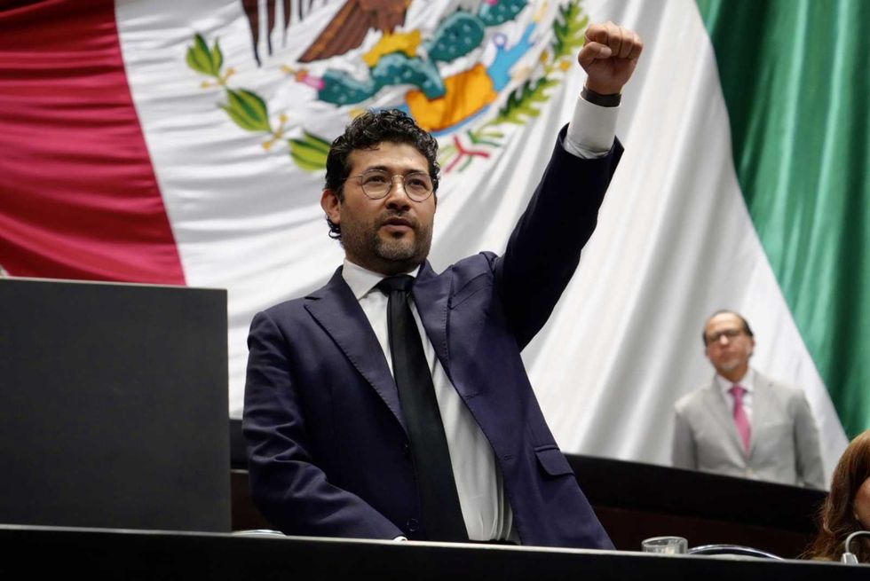 Man raises fist in front of Mexican flag in a formal setting.