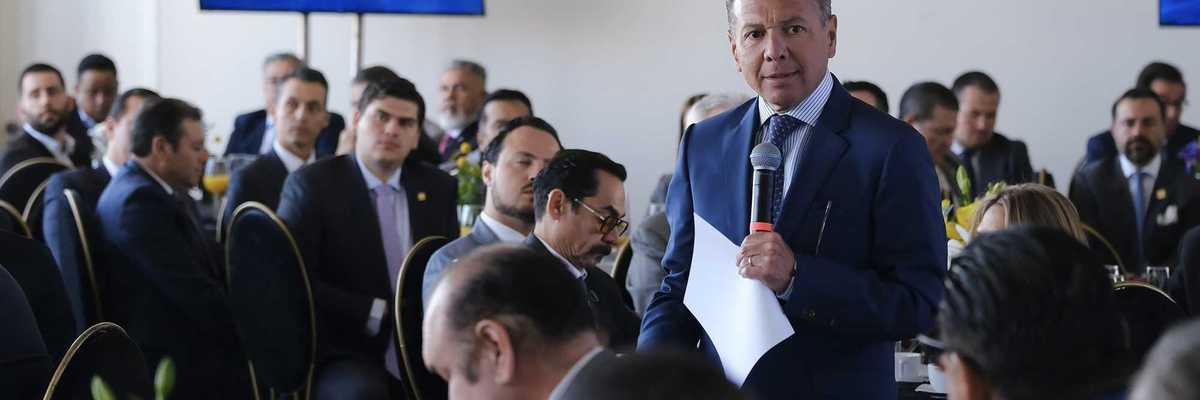 Man speaking at a business meeting with attendees and screens in the background.