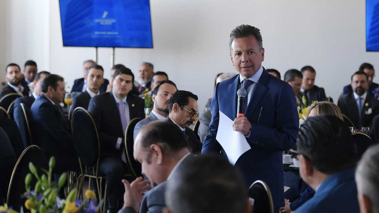 Man speaking at a business meeting with attendees and screens in the background.