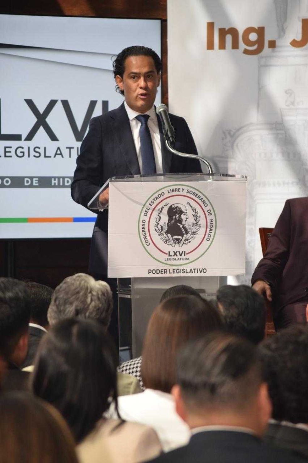 Man speaking at podium during legislative conference, audience listening.