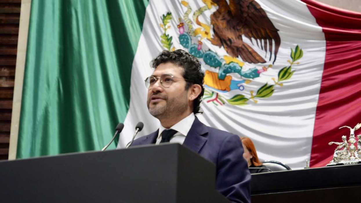 Man speaking at podium with large Mexican flag in background.