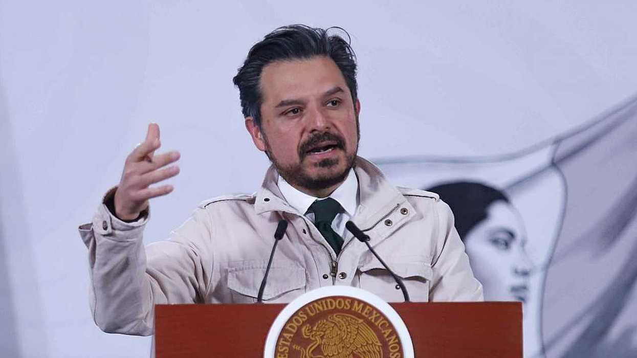 Man speaking at podium with microphones, gesturing with one hand, Mexican emblem visible.