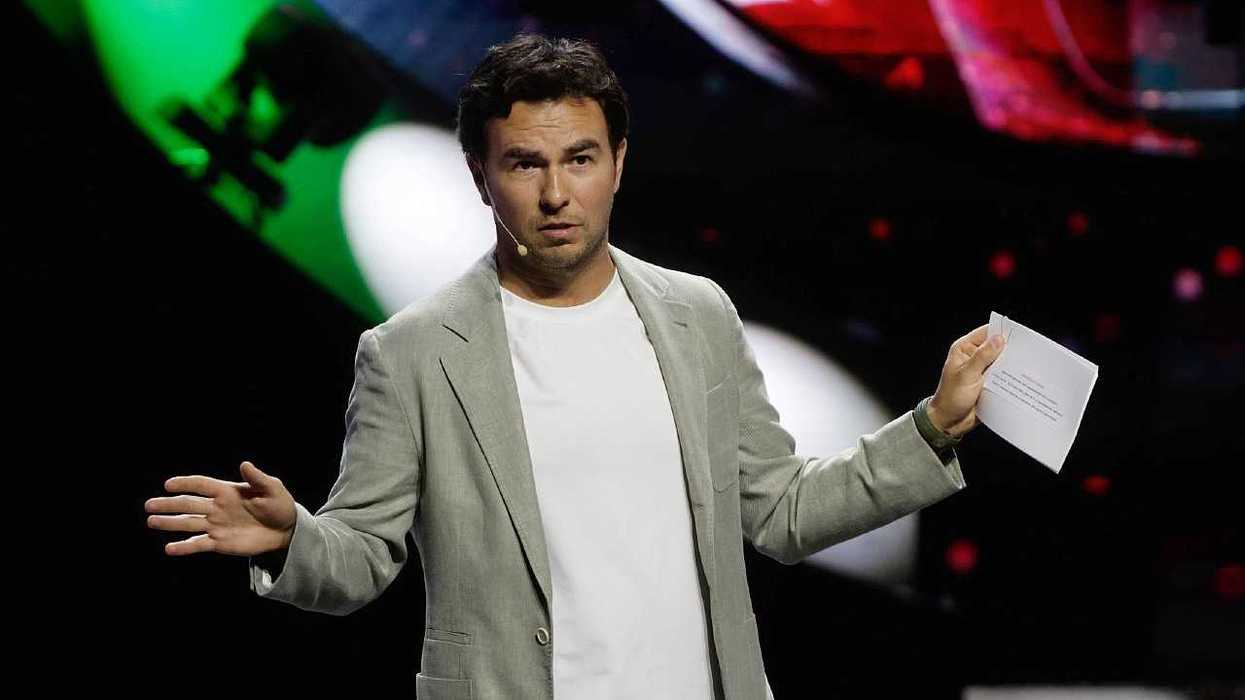 Man speaking on stage, holding papers, green and red lights in background.