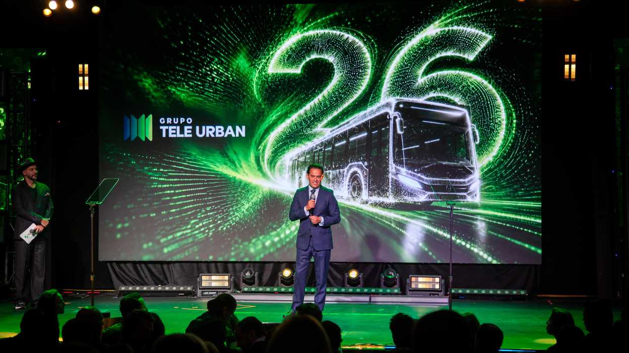 Man speaking on stage with "26" and a bus graphic in green lights behind him.