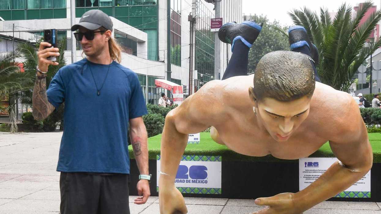 Man takes selfie with large swimmer sculpture in urban setting.