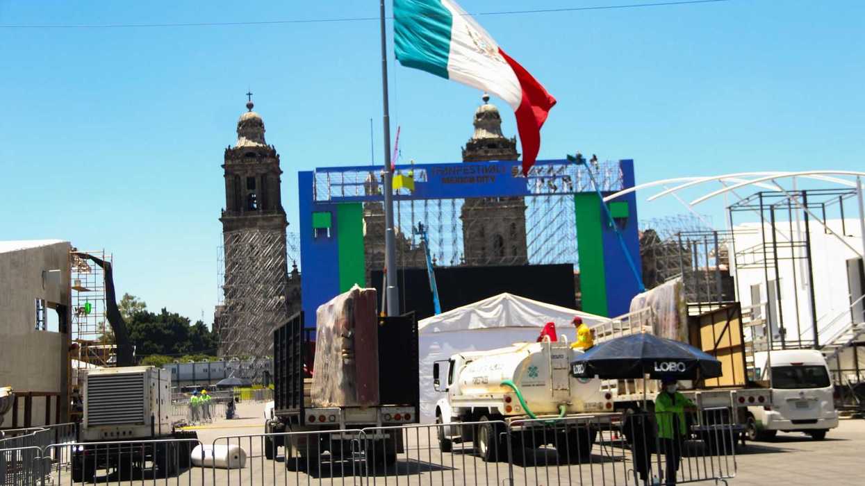 Mexican flag near a stage setup with trucks and scaffolding; church towers in background.