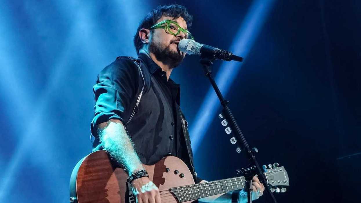 Musician with green glasses playing guitar and singing on stage with blue lights.