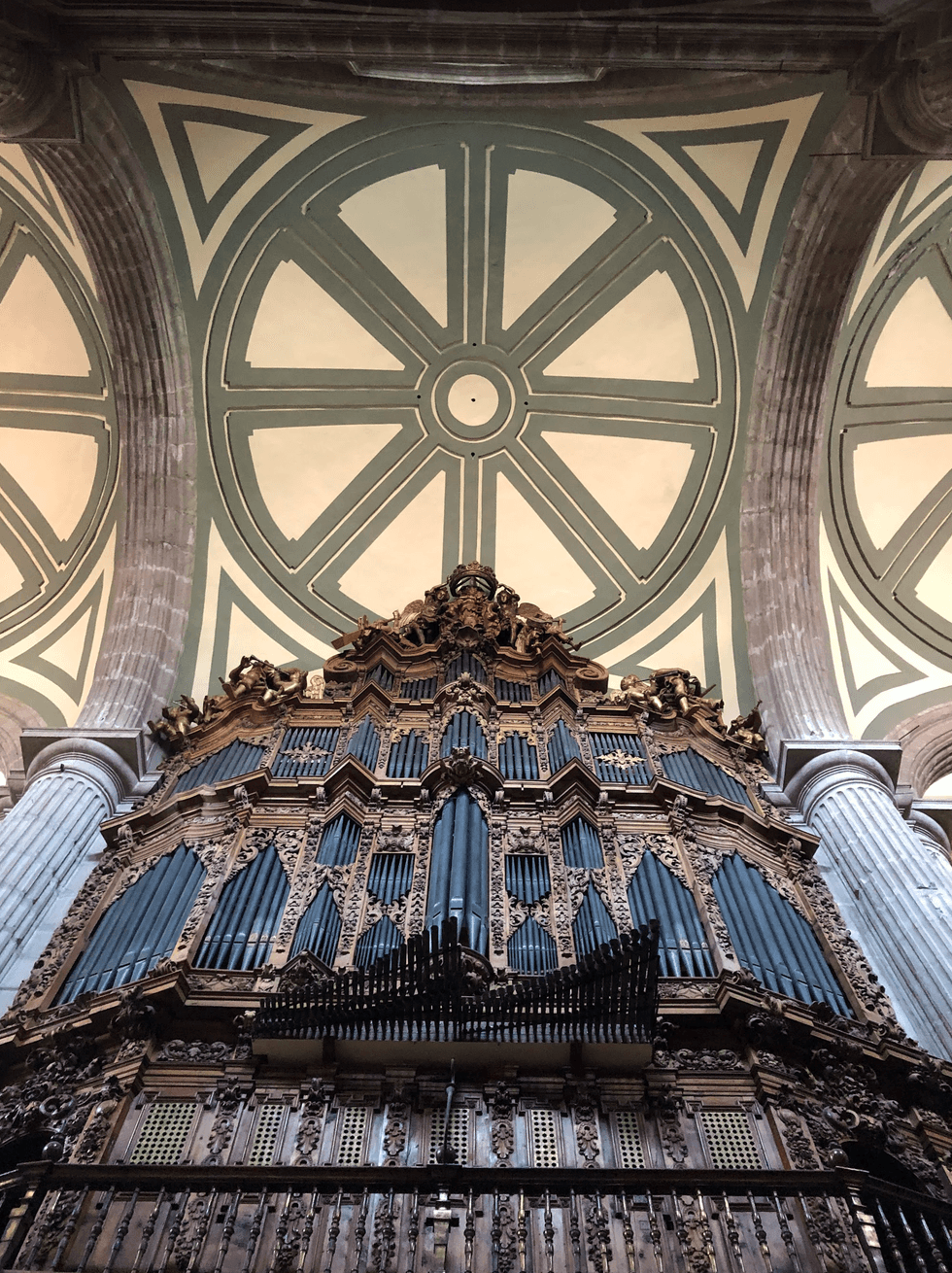 Ornate church organ below a geometric ceiling design with columns.